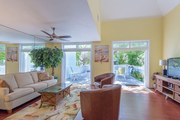 A spacious living room with a sofa, armchairs, a coffee table, and a television. Large windows provide ample natural light and a view of the backyard.