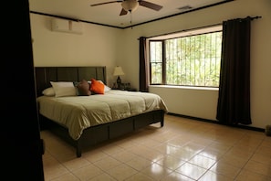 Quarto com cama, ventoinha no teto, janela com cortinas e ar condicionado fixado na parede.