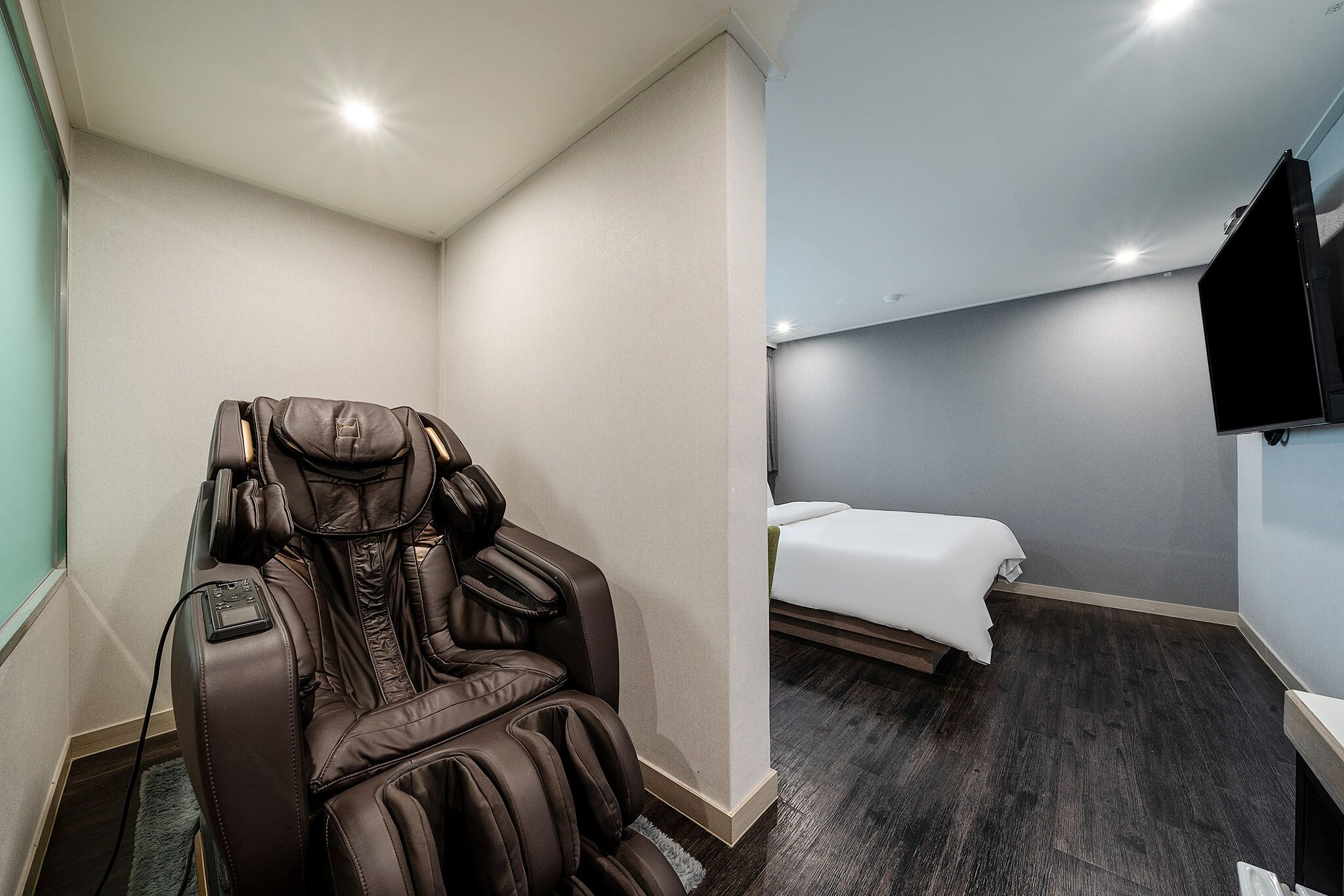 A massage chair in a room with a bed and a flat-screen TV mounted on the wall.