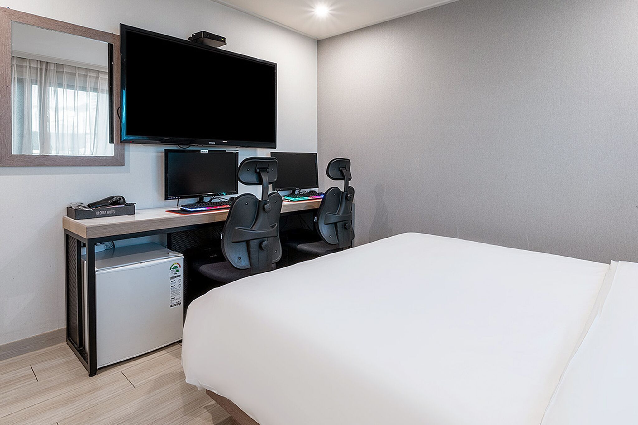 A hotel room with a bed, a desk with two monitors, a keyboard, a mouse, and a small refrigerator.