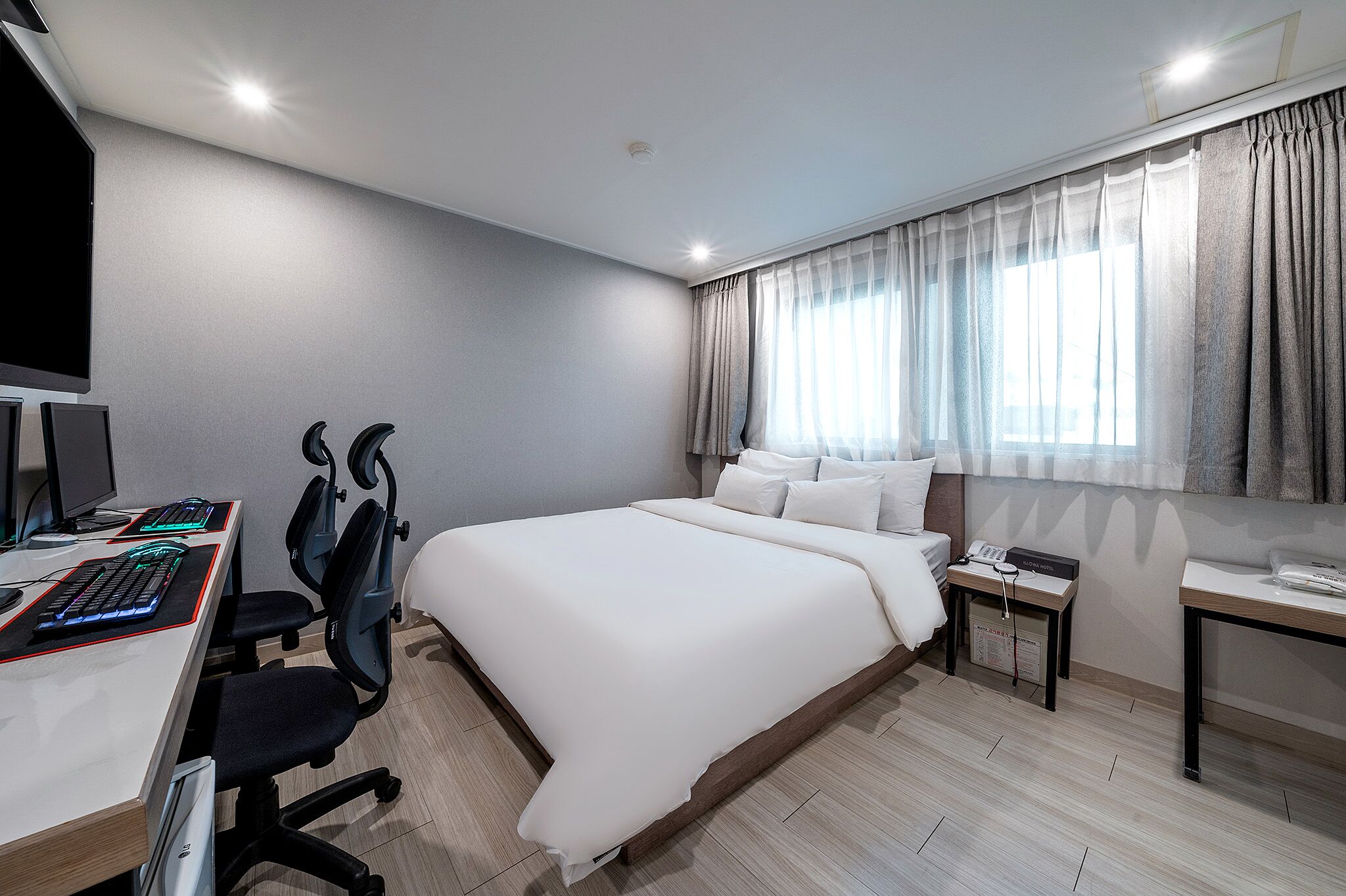 A modern hotel room with a large bed, a desk with a computer and keyboard, a chair, and a bedside table with a lamp.