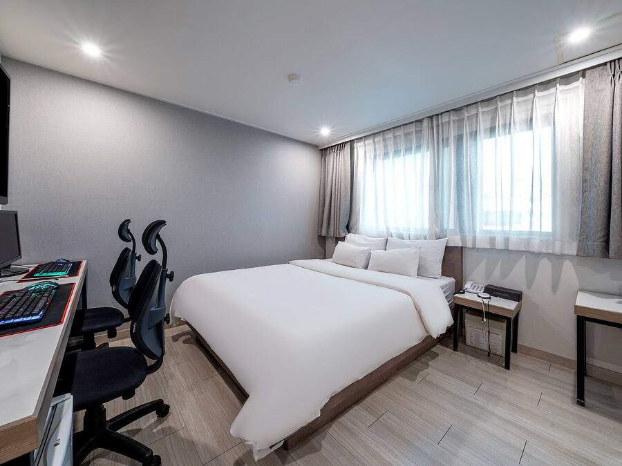 A modern hotel room with a large bed, a desk with a computer and keyboard, a chair, and a bedside table with a lamp.