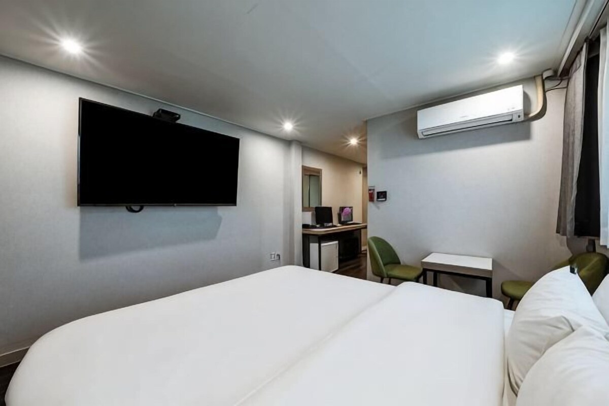 A hotel room with a large bed, a TV mounted on the wall, a desk with a chair, and an air conditioning unit.