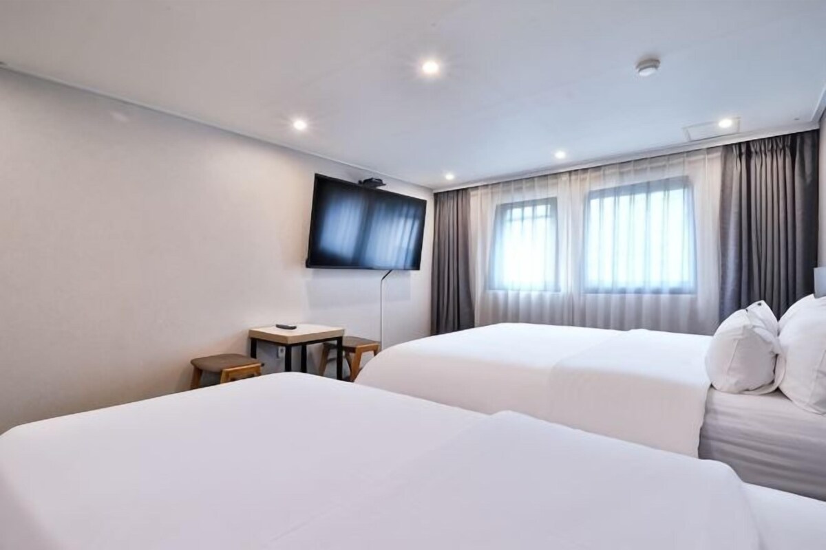 A hotel room with two beds, a flat-screen TV, a small table, and a window with curtains.