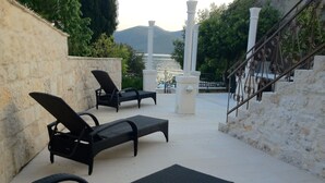 A sunlit outdoor seating area with lounge chairs and a stone wall, overlooking a body of water and hills. - Seafront Historic Villa | Private pool, kayaks and bikes (Viganj)