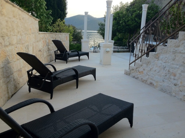A sunlit outdoor seating area with lounge chairs and a stone wall, overlooking a body of water and hills.