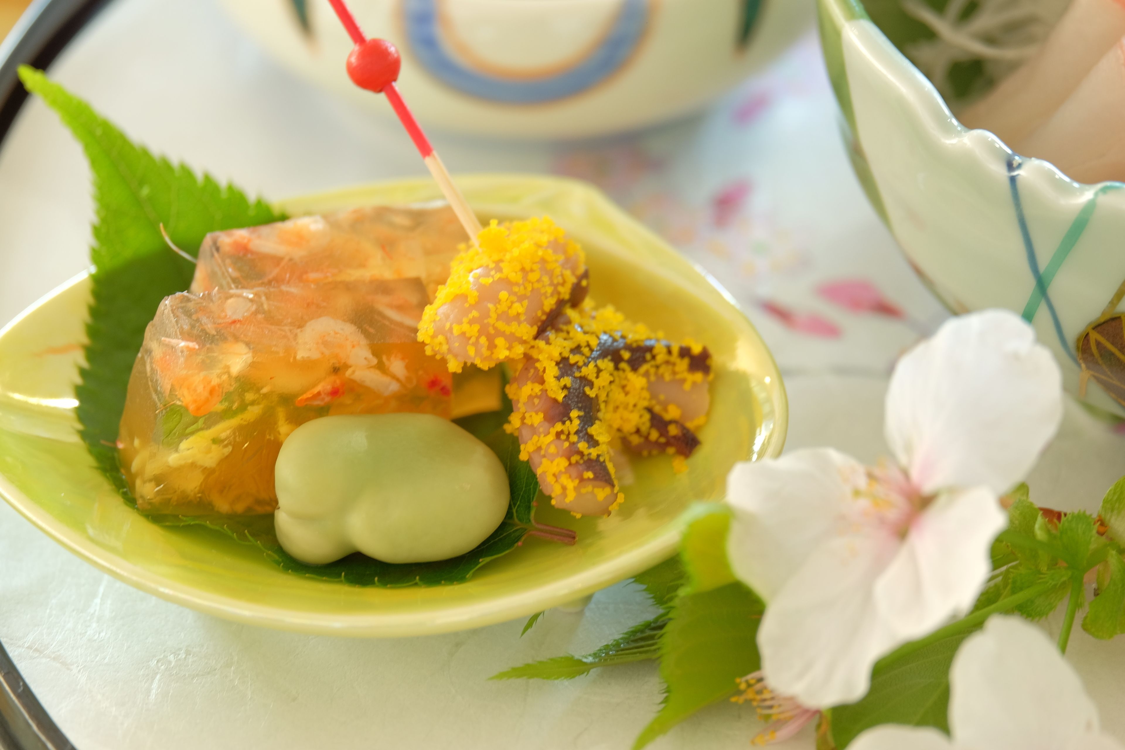 A plate with various food items, including a translucent jelly-like dish, a yellow crumbly item, and a green oval-shaped item, all on a green leaf.