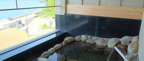 A hot spring area with a stone border, a wooden bench, and a view of the sea through a window.