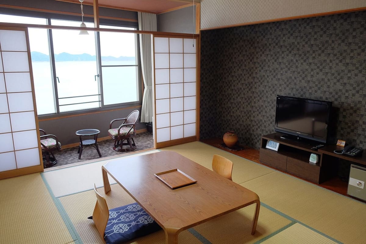 Una habitación japonesa tradicional con suelo de tatami, una mesa baja con cojines, televisión y vista al mar a través de puertas shoji.