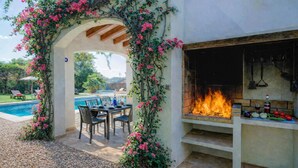 Eine Terrasse im Freien mit Pool, einem Essplatz und einem Kamin aus Stein mit Feuer.
