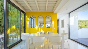 A modern dining area with a white table and chairs, a yellow accent wall, and large windows offering a view of greenery. - Designer Alcanada by Balearic Villas (Alcúdia)