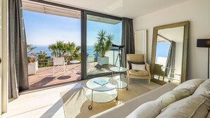 A modern hotel room with a large sliding glass door leading to a balcony with a view of the sea. There is a sofa, a small table, and a chair on the balcony. - Designer Alcanada by Balearic Villas (Alcúdia)