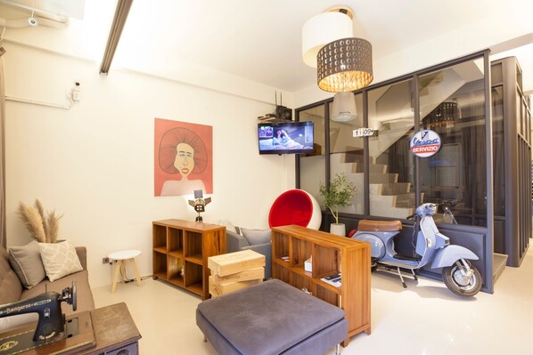 Una sala moderna con una máquina de coser antigua, un televisor, un sillón rojo, un patinete y un ventanal con un letrero.