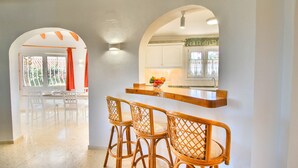 Een moderne keuken met witte kastjes, een houten bar en een fruitschaal. Drie rieten stoelen staan voor de bar.