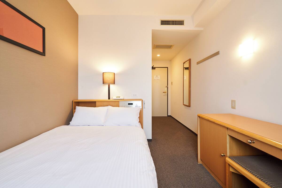 A hotel room with a bed, bedside table, lamp, and a door leading to another room.