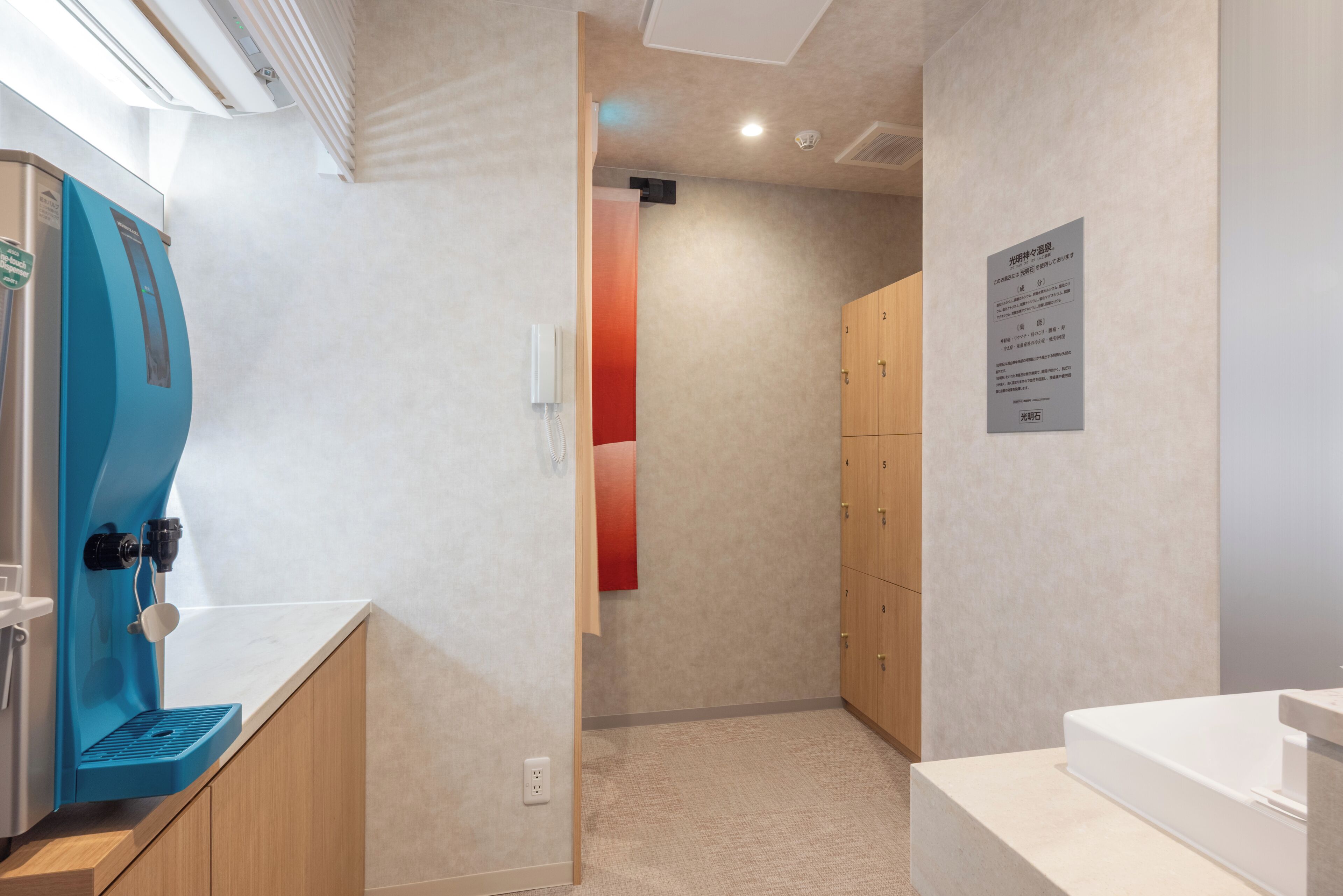 A hotel room with a blue water dispenser, wooden cabinets, a white sink, and a beige carpet.