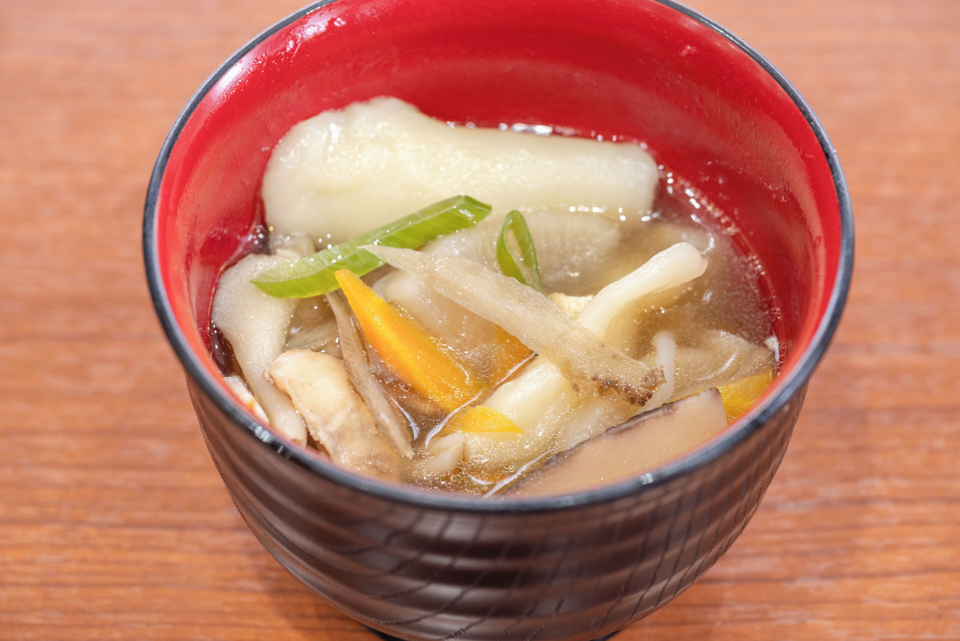 A bowl of clear soup with vegetables and a white item, possibly tofu, in a red bowl on a wooden surface.