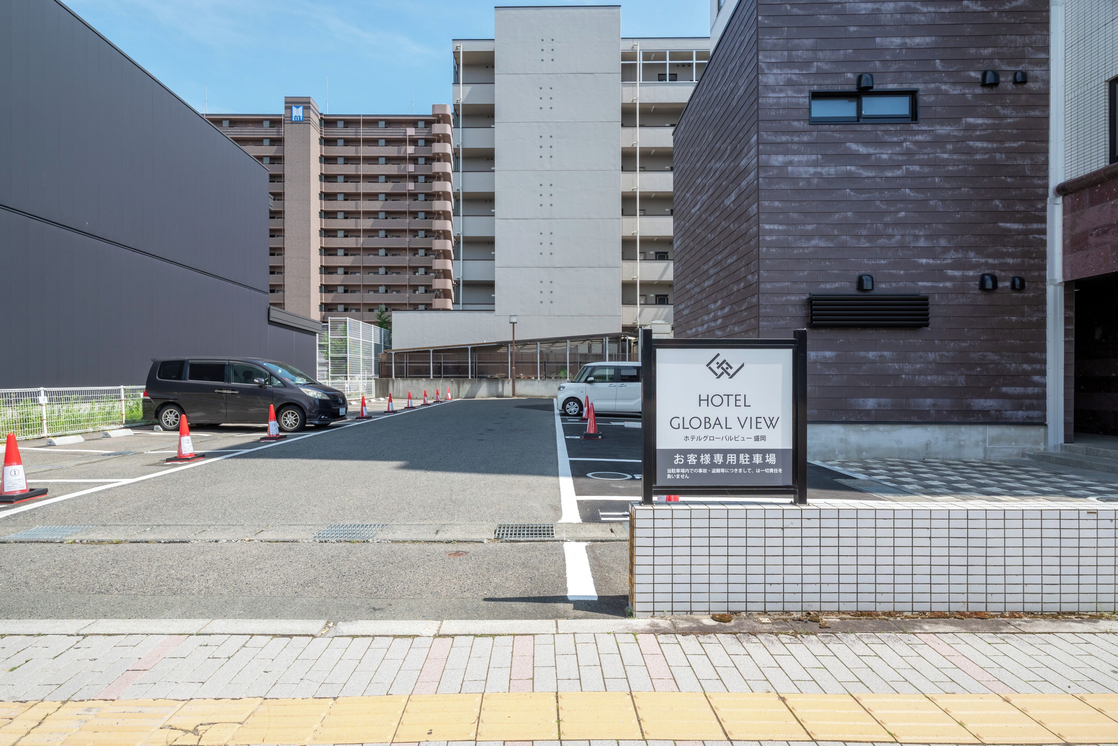 A hotel entrance with a sign reading 'HOTEL GLOBAL VIEW,' a parking area with a few cars, and a modern building with a tiled facade.