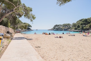 A sandy beach with a concrete path, rocky outcrops, and dense trees.
