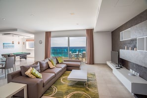 Una sala moderna con sofá, mesa de centro y televisor. Se observa una mesa de billar al fondo y vistas al mar a través de amplias ventanas.