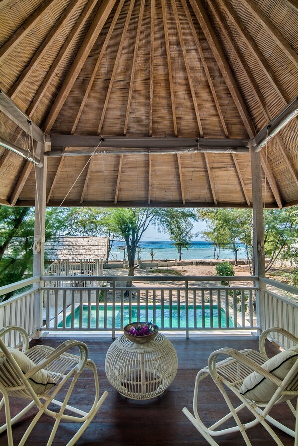 A covered outdoor area with a wooden ceiling, white wicker chairs, a wicker table, and a view of a swimming pool and beach. - Villa Coral Flora (Gili Trawangan)