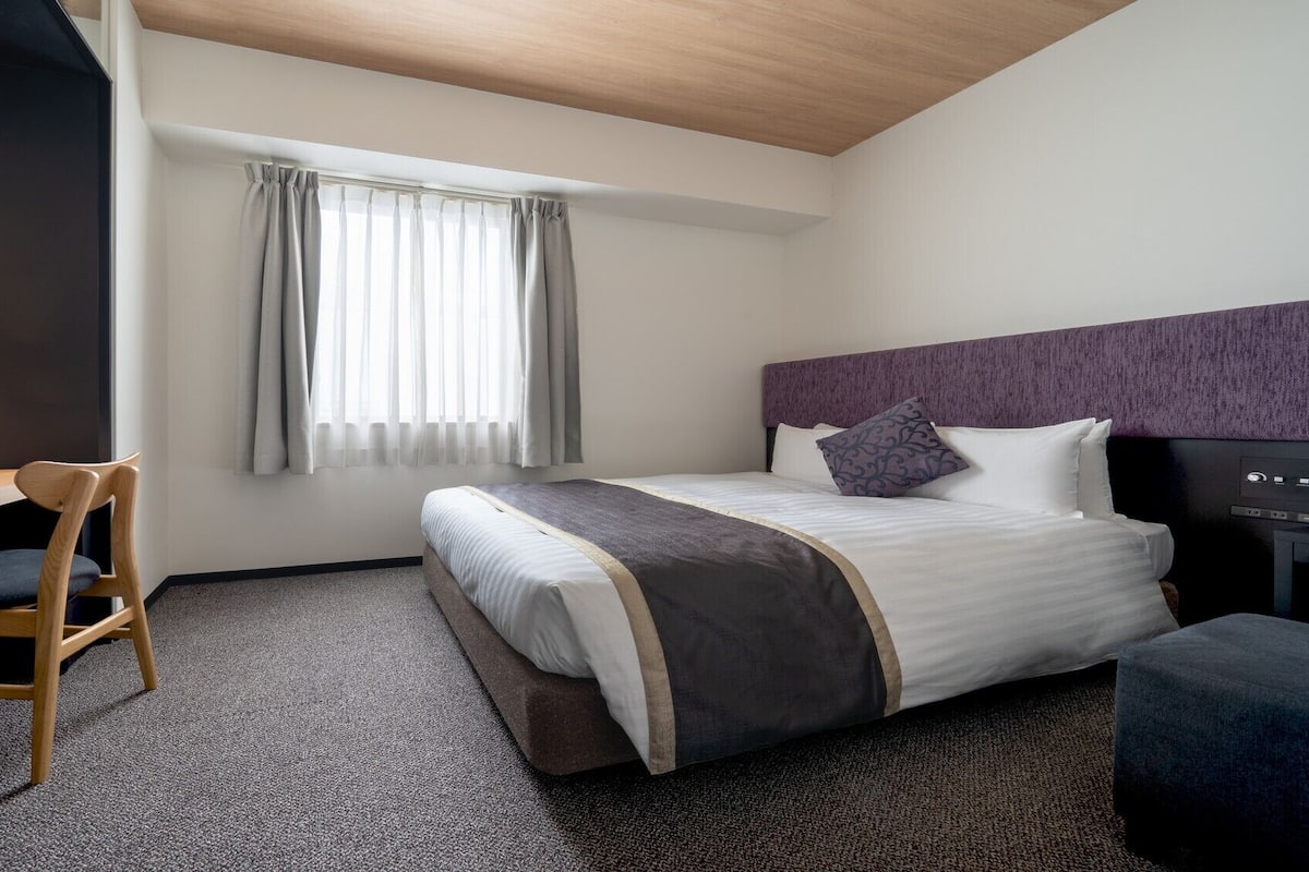 Ein Hotelzimmer mit Bett, Schreibtisch, Stuhl, Fernseher und einem Fenster mit Vorhängen.