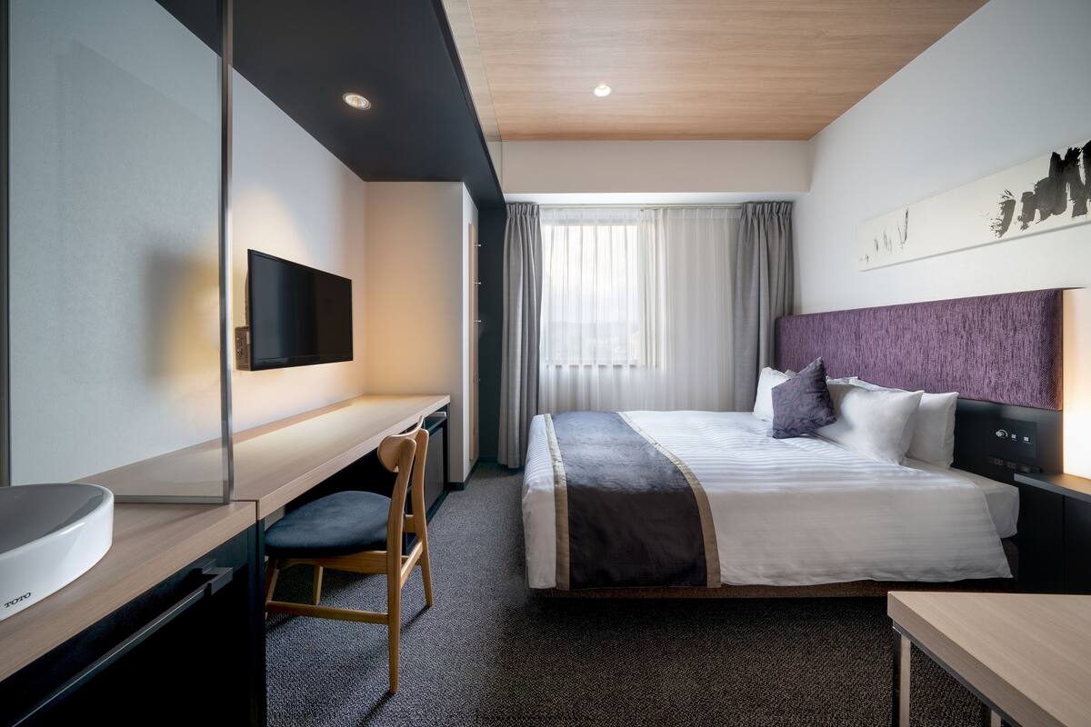 A modern hotel room with a bed, a desk, a chair, a TV, and a window with curtains.