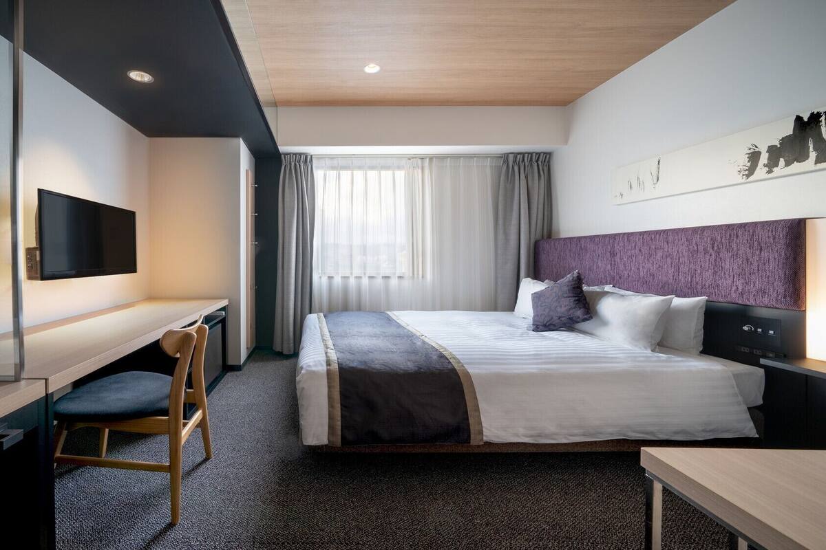 A hotel room with a bed, a desk, a chair, a TV, and a window with curtains.