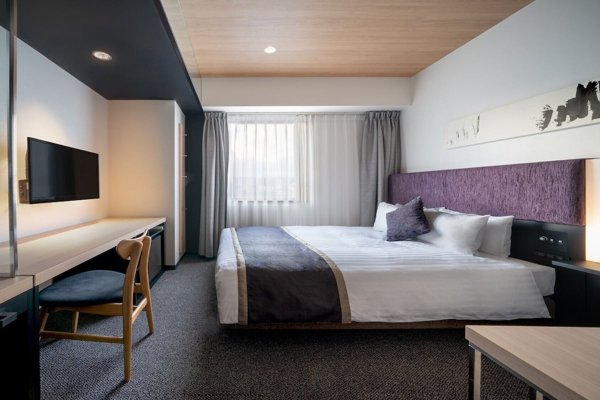 Ein Hotelzimmer mit Bett, Schreibtisch, Stuhl, Fernseher und einem Fenster mit Vorhängen.