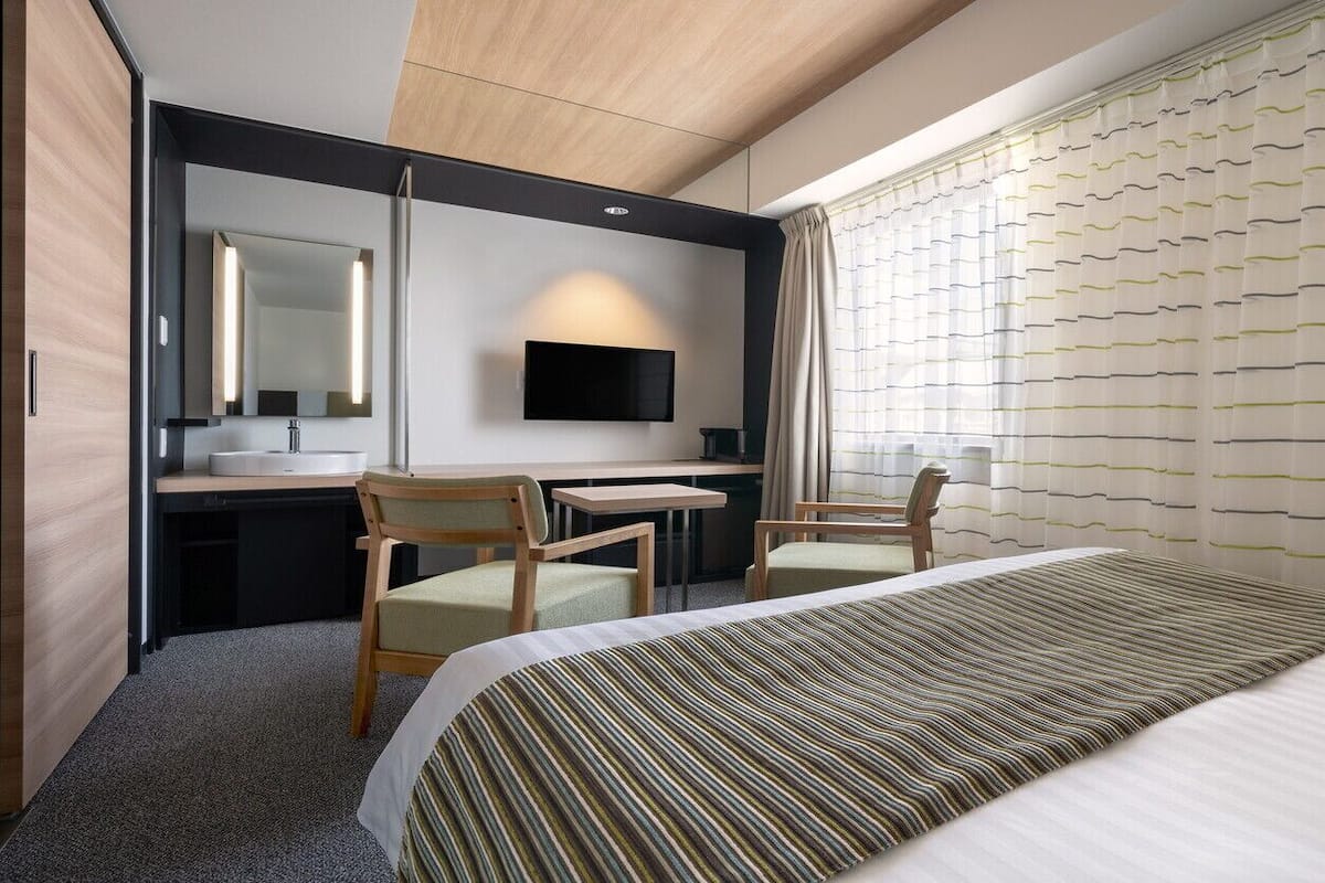 A hotel room with a bed, a desk with a chair, a television, and a window with curtains.