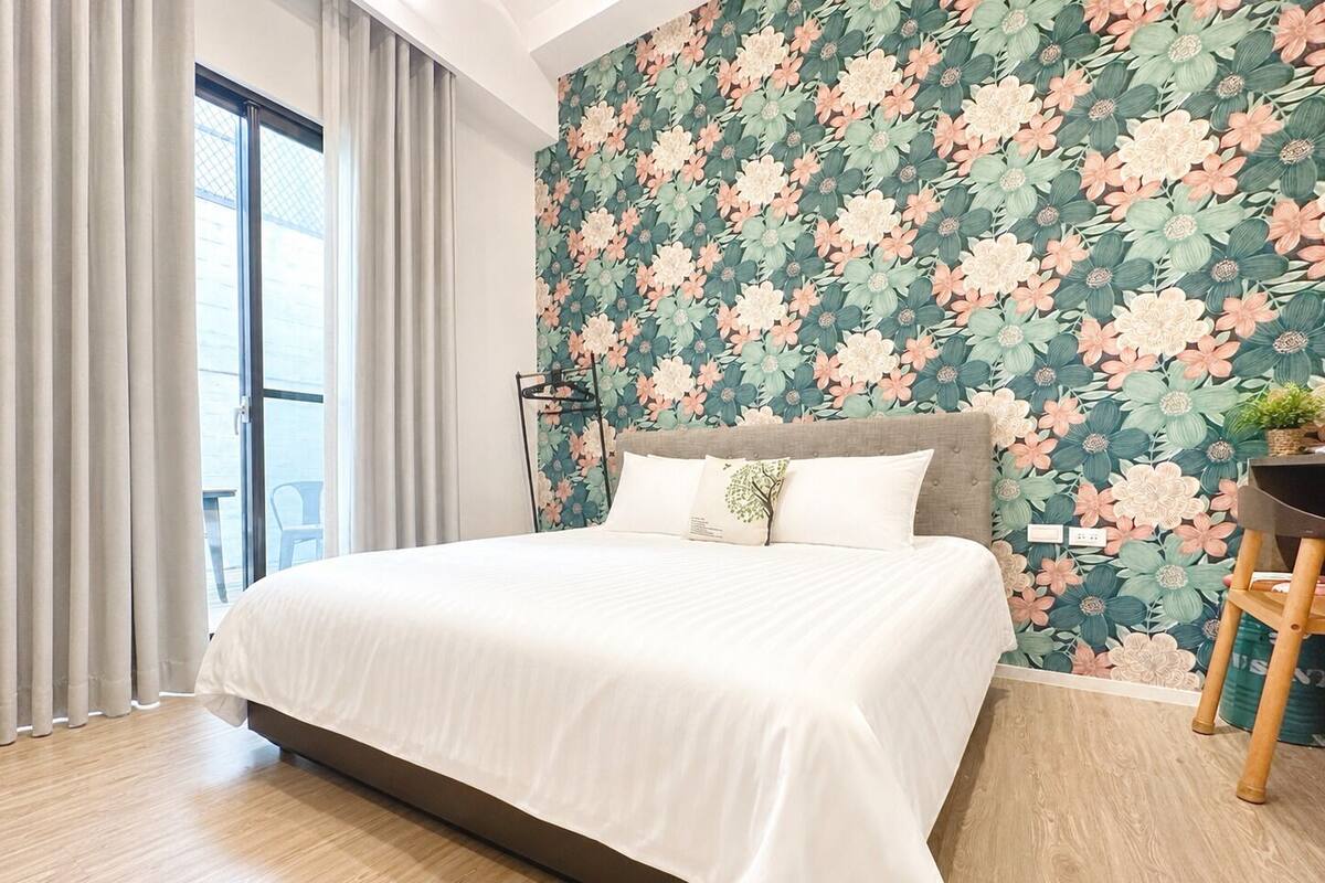 Quarto moderno com cama grande, cabeceira, janela com cortinas e papel de parede floral.