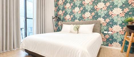 A modern bedroom with a large bed, a headboard, a window with curtains, and a floral wallpaper.