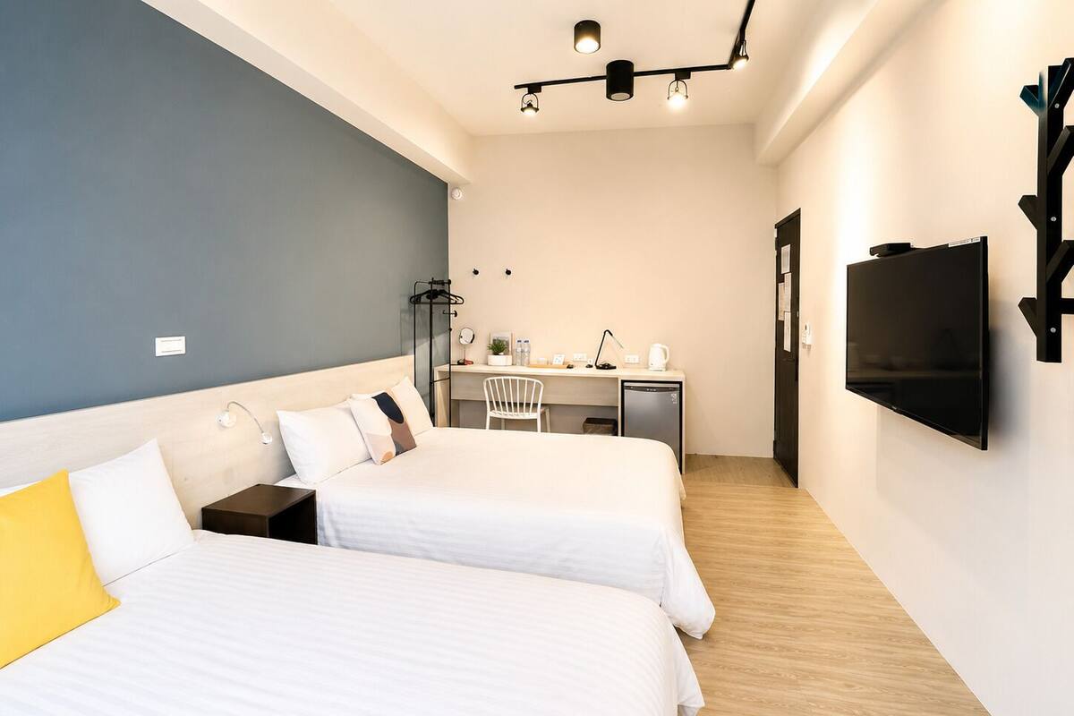 Quarto de hotel com duas camas, uma escrivaninha, uma cadeira, uma televisão e um guarda-roupa.