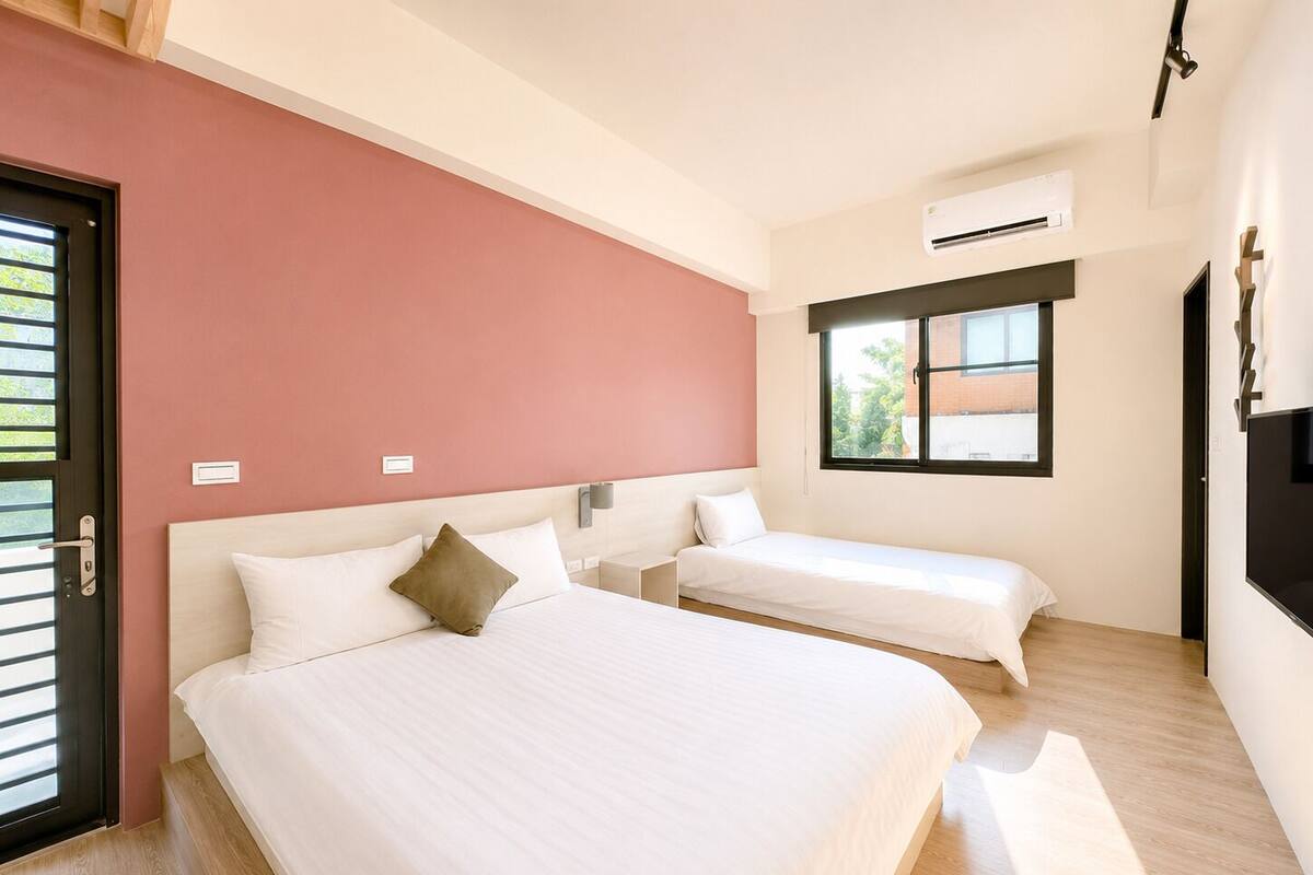 Quarto de hotel com duas camas, uma parede de destaque vermelha, uma janela com vista e um aparelho de ar condicionado.