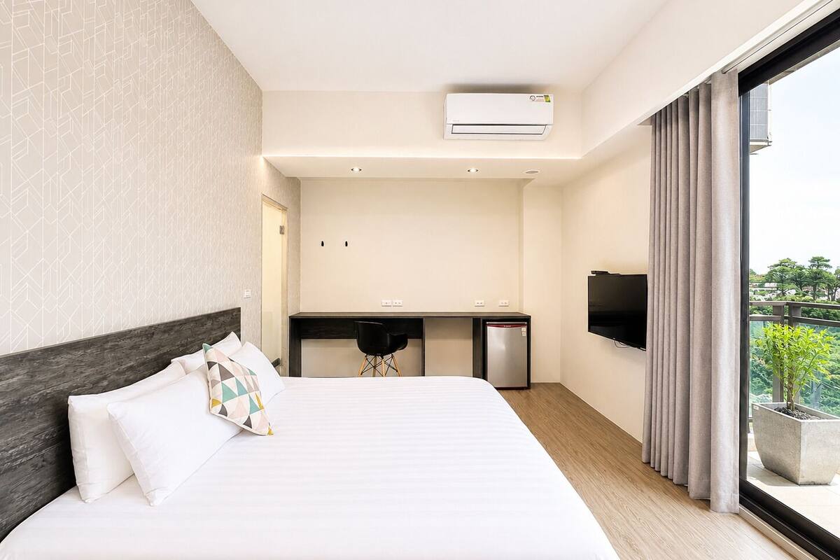 Quarto de hotel com cama, mesa de trabalho, televisão e varanda com planta.