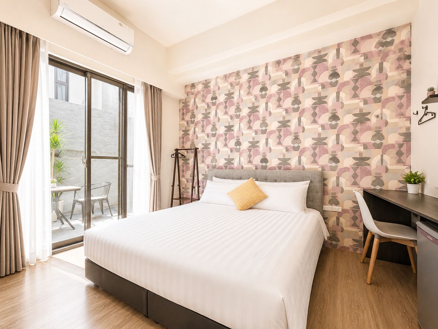 Habitación de hotel con cama, escritorio, silla, ventana y pared con diseño geométrico.