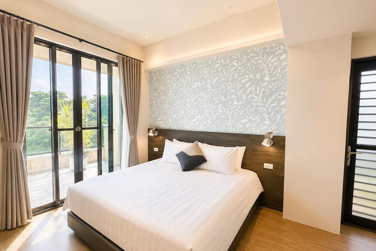 Quarto de hotel moderno com uma cama grande, uma varanda com cortinas, parede estampada e piso de madeira.