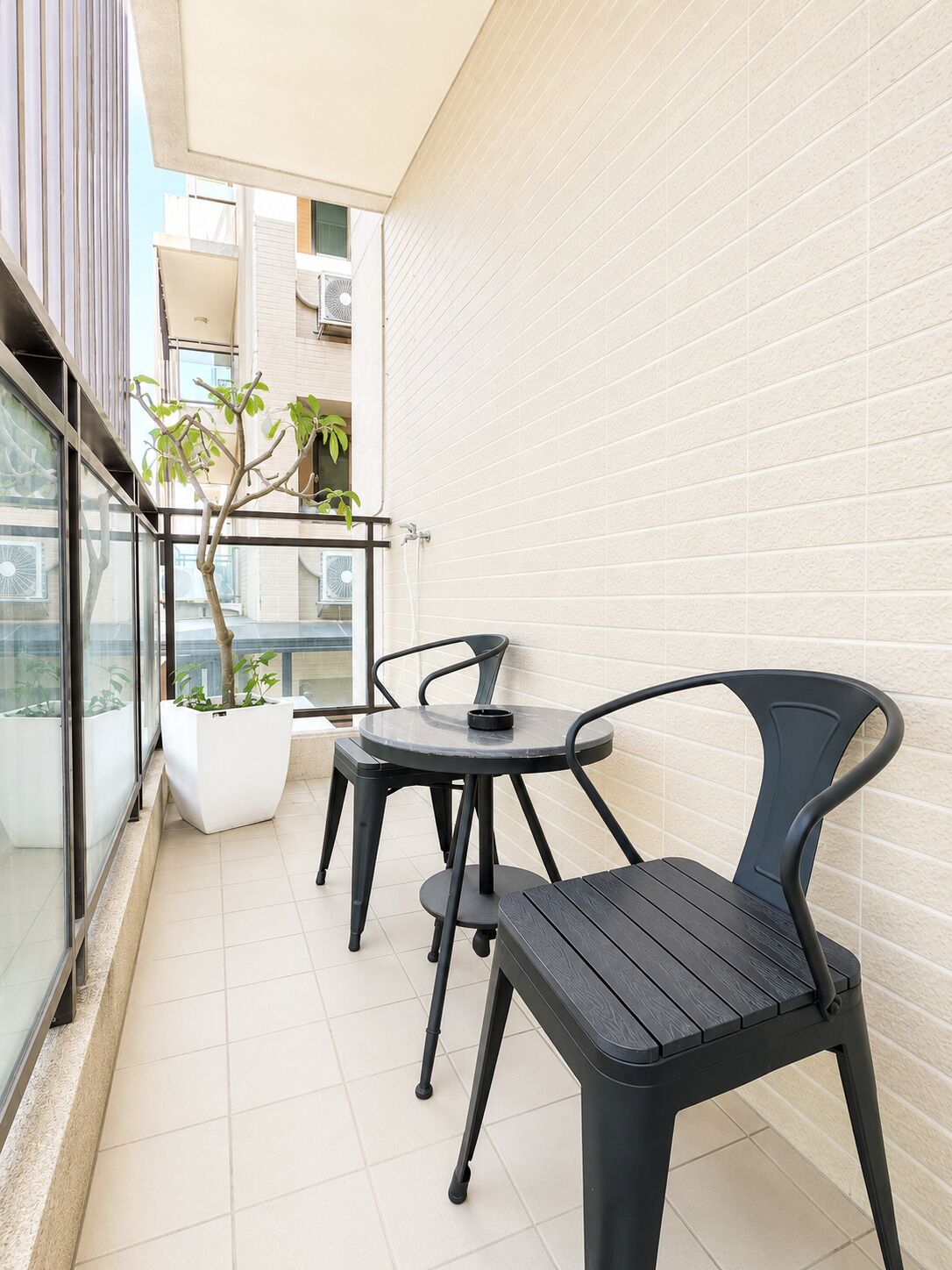 En balkon med et lille bord, to stole, en potteplante og udsigt til en bygning.