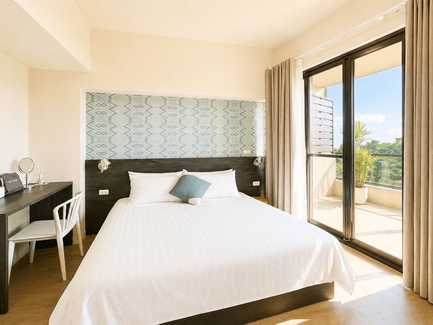Habitación de hotel con cama, escritorio, silla, ventana grande con cortinas y pared decorada.