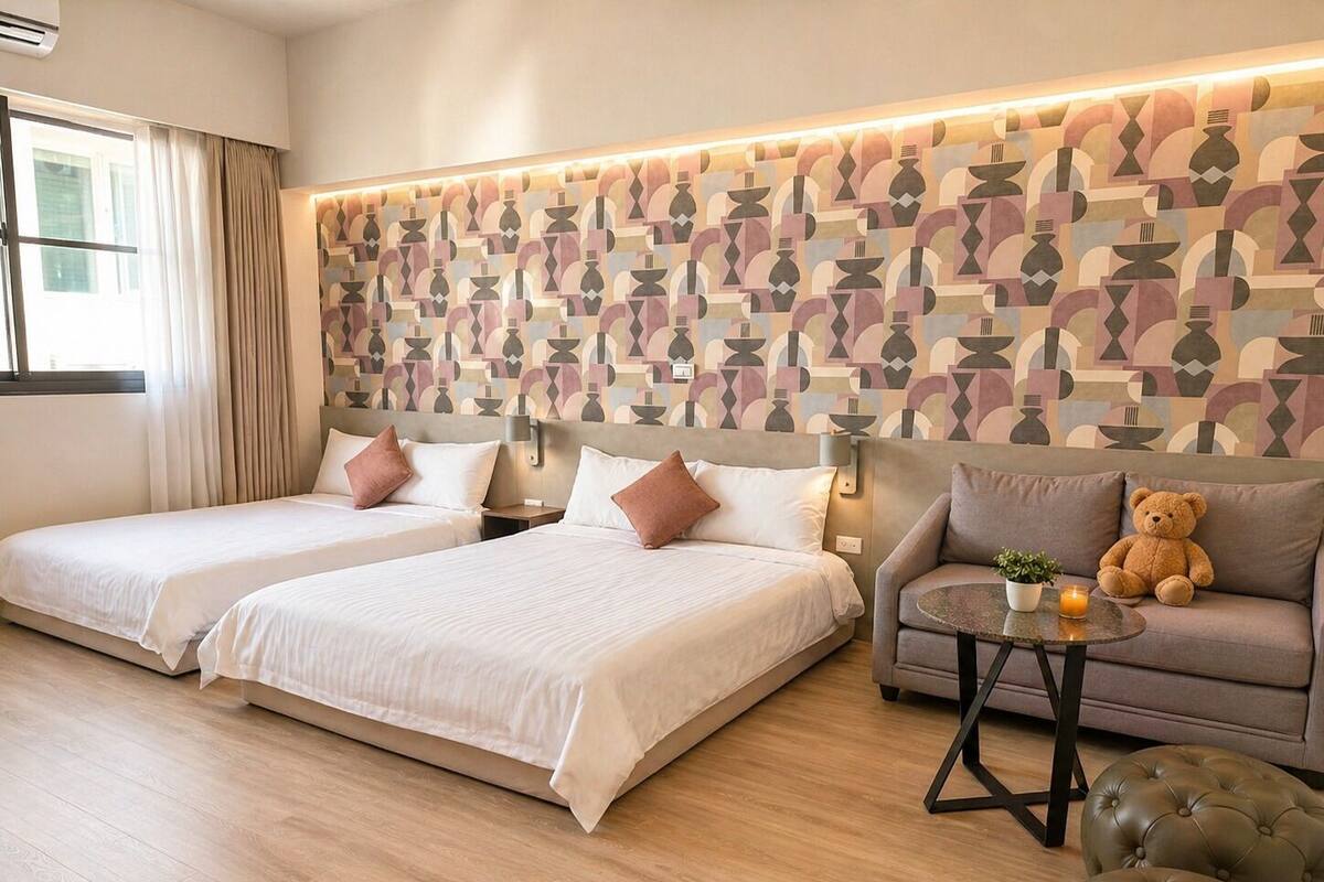 Quarto de hotel com duas camas, um sofá, um urso de pelúcia, uma mesinha com uma planta e uma vela, e parede com padrão geométrico.