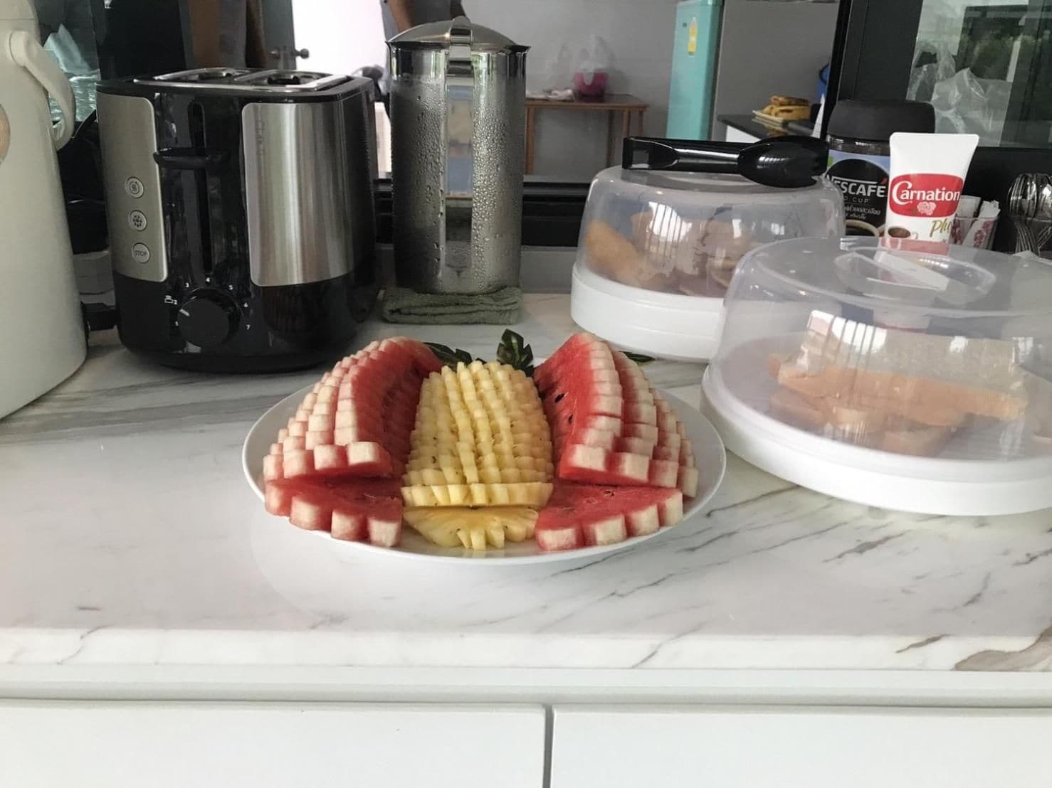 Een keukentafel met een bord met gesneden fruit, een koffiezetapparaat, een waterkoker en een koffiekop.