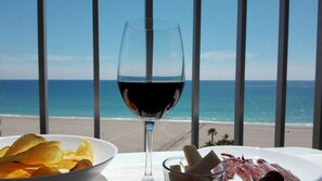 Un tavolo con un bicchiere di vino rosso, una ciotola di patatine e un piccolo piatto di pancetta, con vista su un balcone che si affaccia sulla spiaggia e sull'oceano.