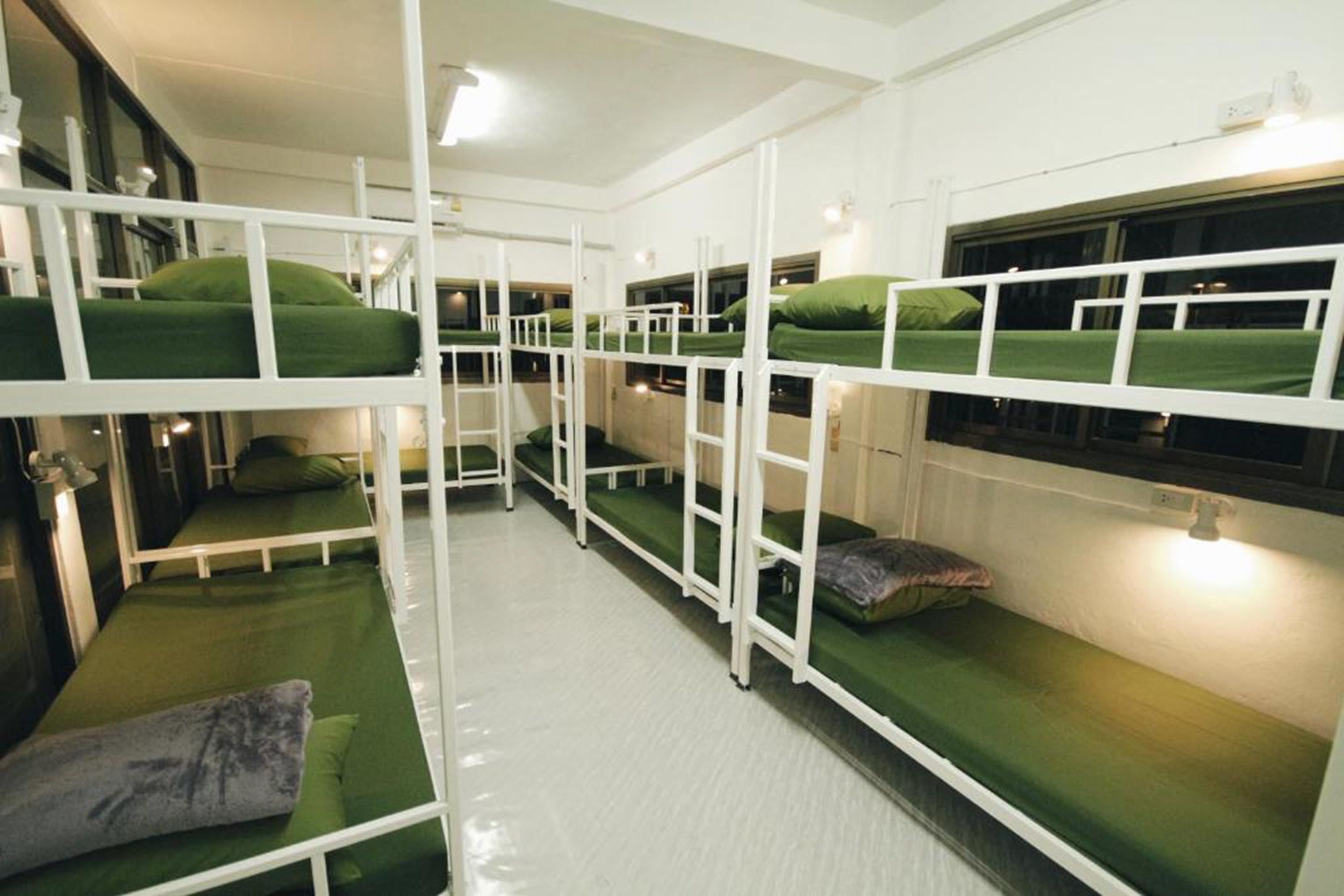 A dormitory-style room with bunk beds, each with a green mattress and pillow, white metal railings, and a white floor.
