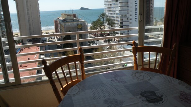Un balcon avec une table et des chaises, donnant sur un paysage urbain côtier avec des bâtiments et offrant une vue dégagée sur la mer.