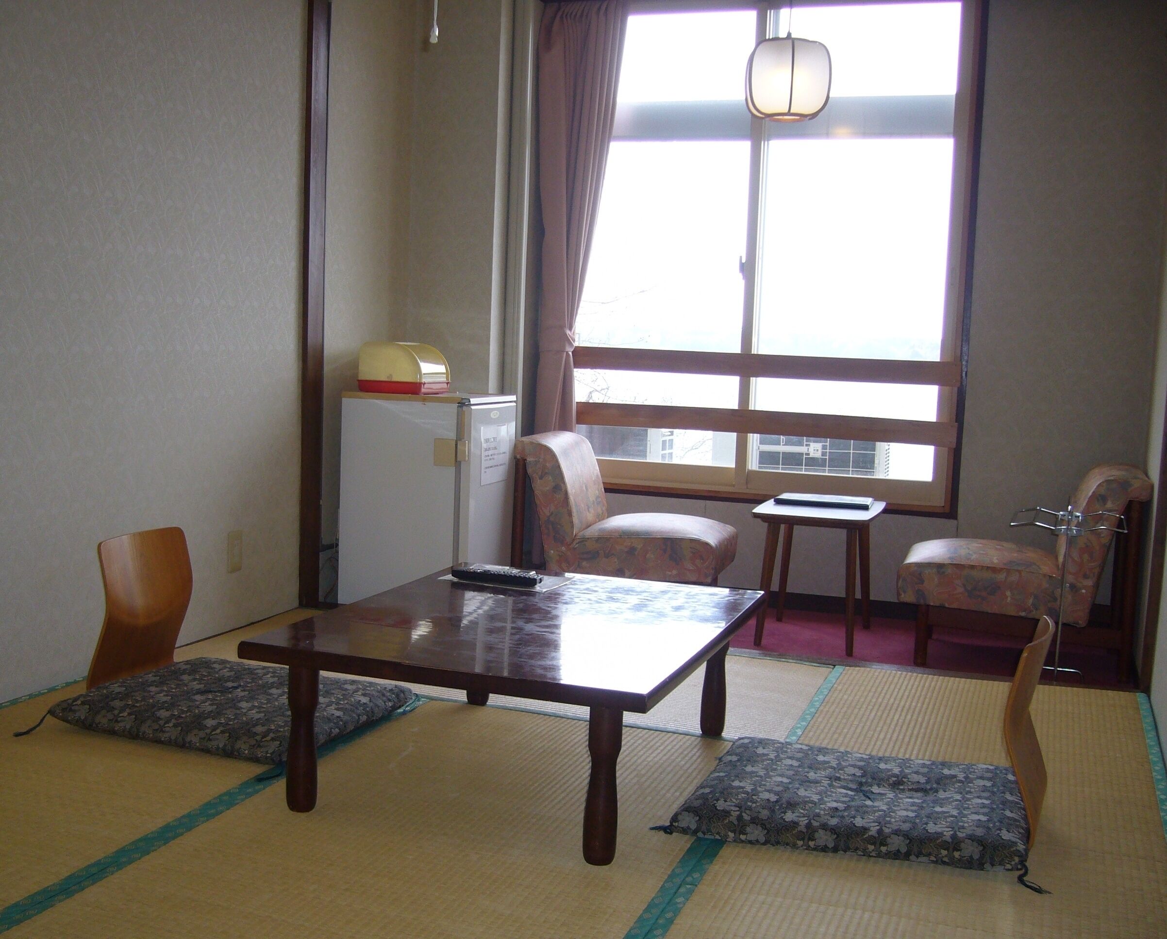 Ett traditionellt japanskt rum med tatamimattor på golvet, ett lågt bord, två stolar, ett litet bord och ett kylskåp.
