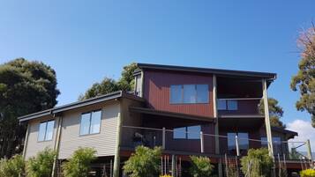 A two-story house with a balcony on the upper floor, surrounded by trees and a stone wall.