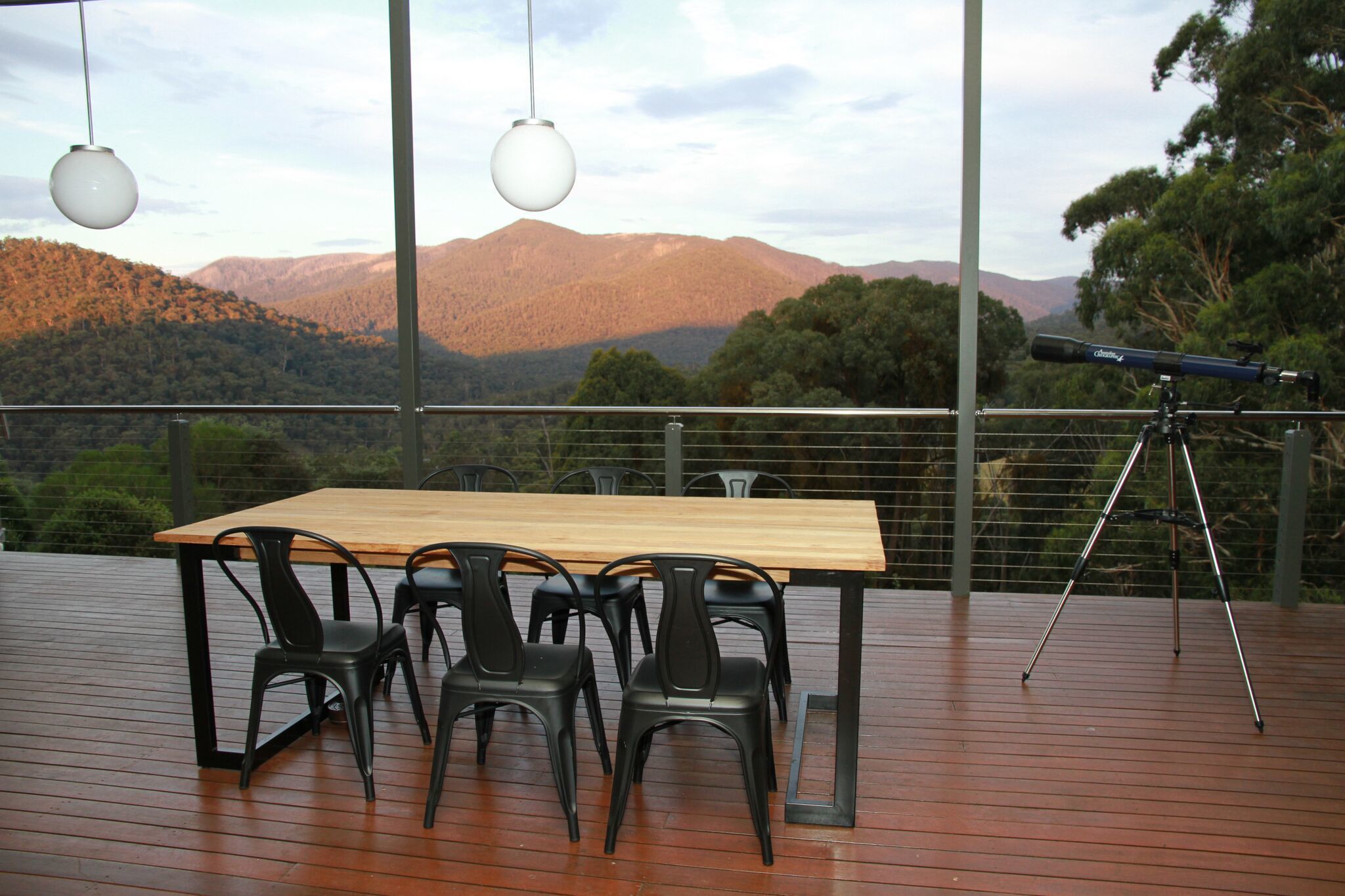 Una terraza de madera con una mesa y sillas, un telescopio, y vistas a montañas y árboles.