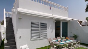 A white building with a balcony, a dining area with a table and chairs, and a staircase leading to the upper floor.