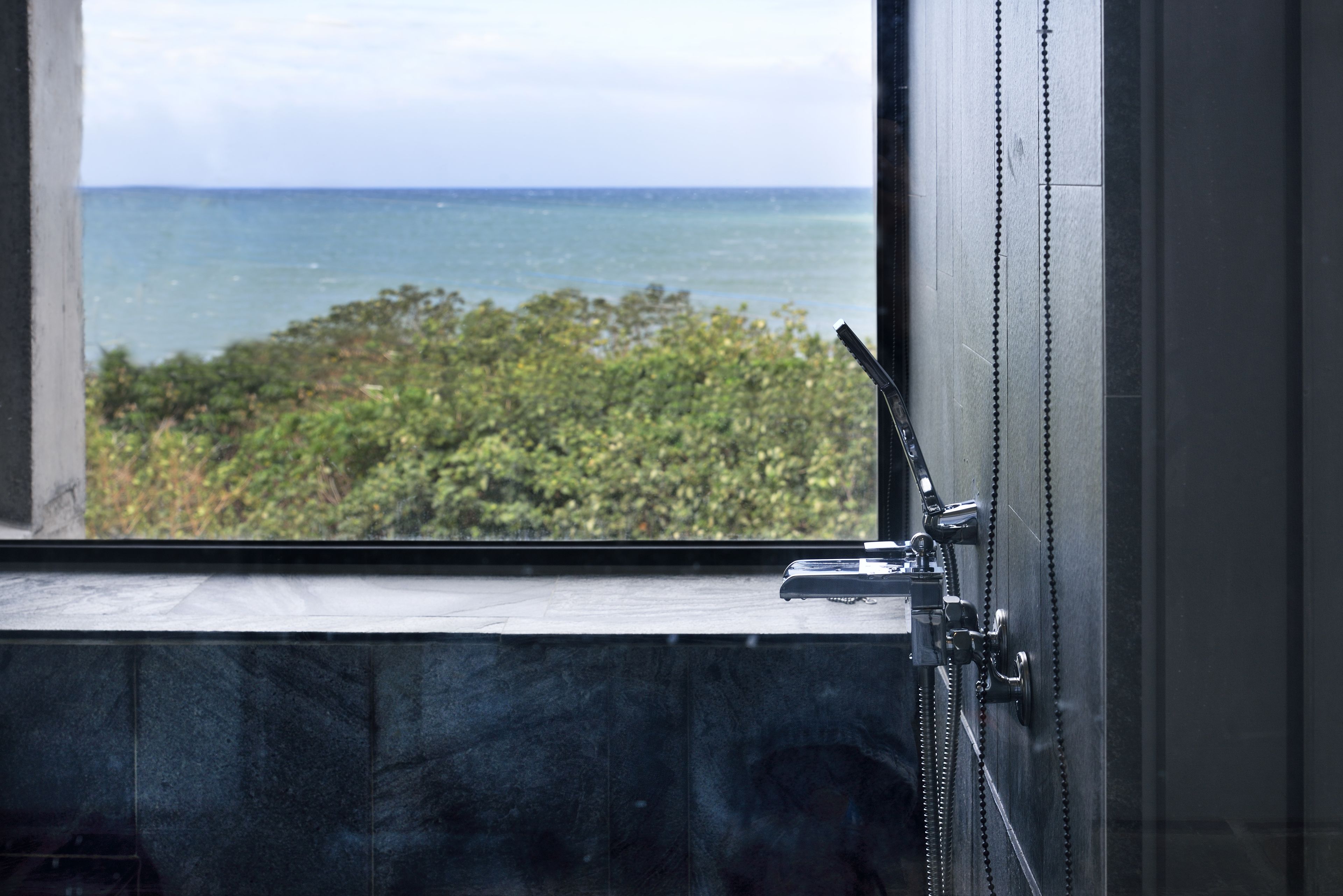 Un baño con vista al océano a través de una ventana, cabezal de ducha y suelo de baldosas.