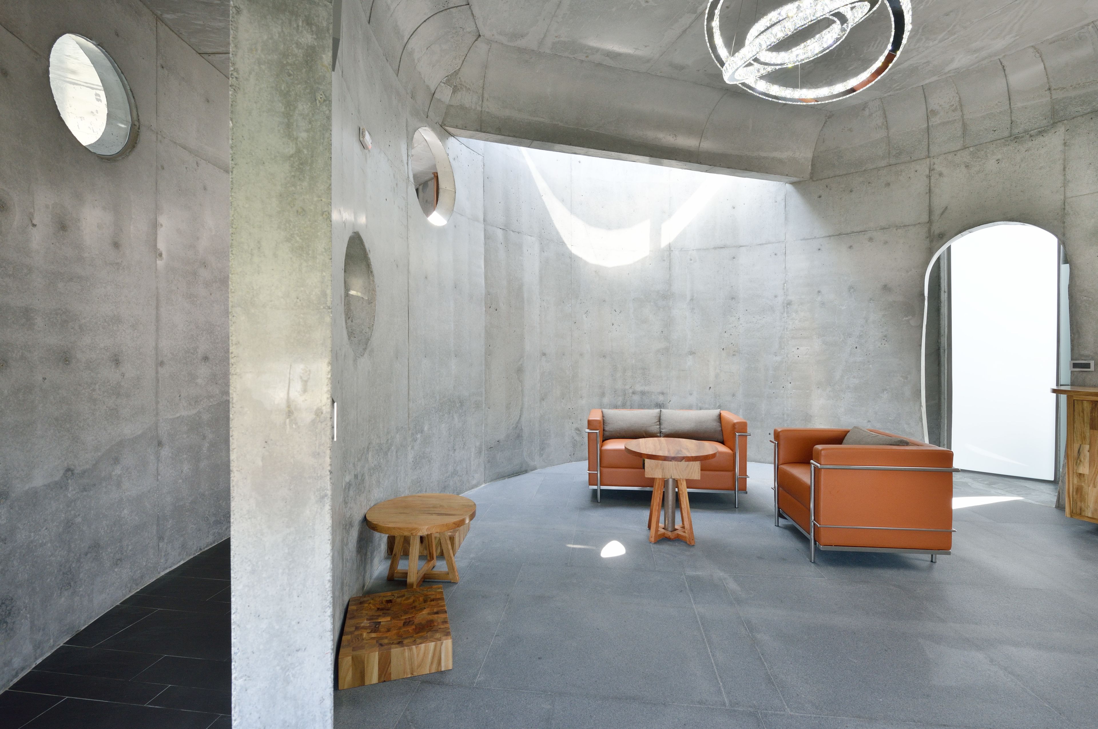 A modern interior with a concrete wall, a round wooden table, two orange sofas, and a round wooden stool.